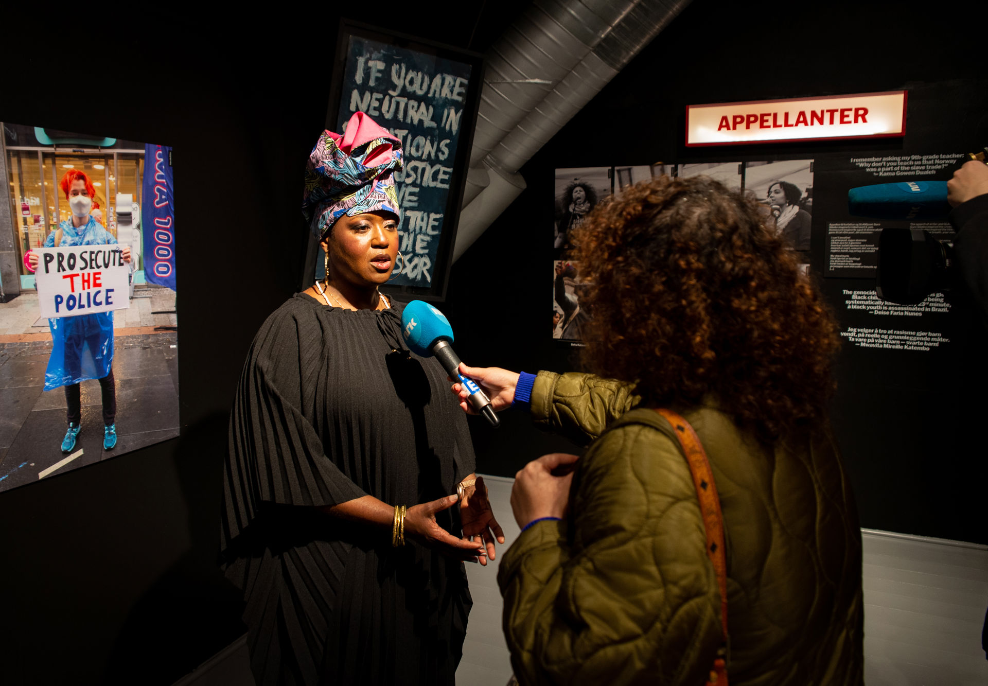 Journalist fra NRK holder en blå mikrofon foran prosjektansvarlig Michelle Tisdel inne i utstillingen Din pust, din stemme på Bymuseet i Frognerparken.