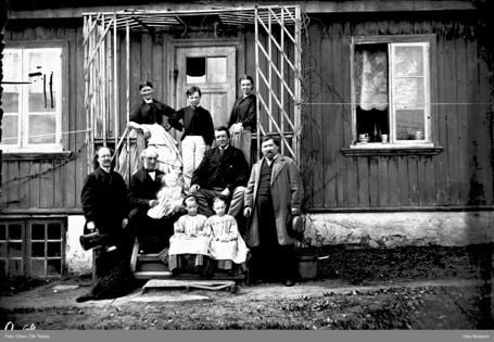 sorthvitt av voksne og barn samlet på en trapp foran et gammelt litt slitent trehus