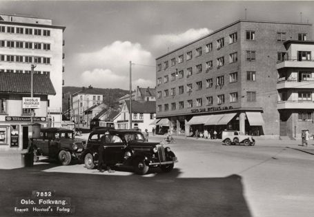 sort-hvitt av to gamle biler på et torv med bolig- og butikkbebyggelse rundt