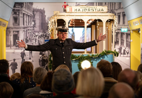 Mann i trikkeuniform med hvit lue og sort dress står med armene ut foran en blå trikk i en utstilling