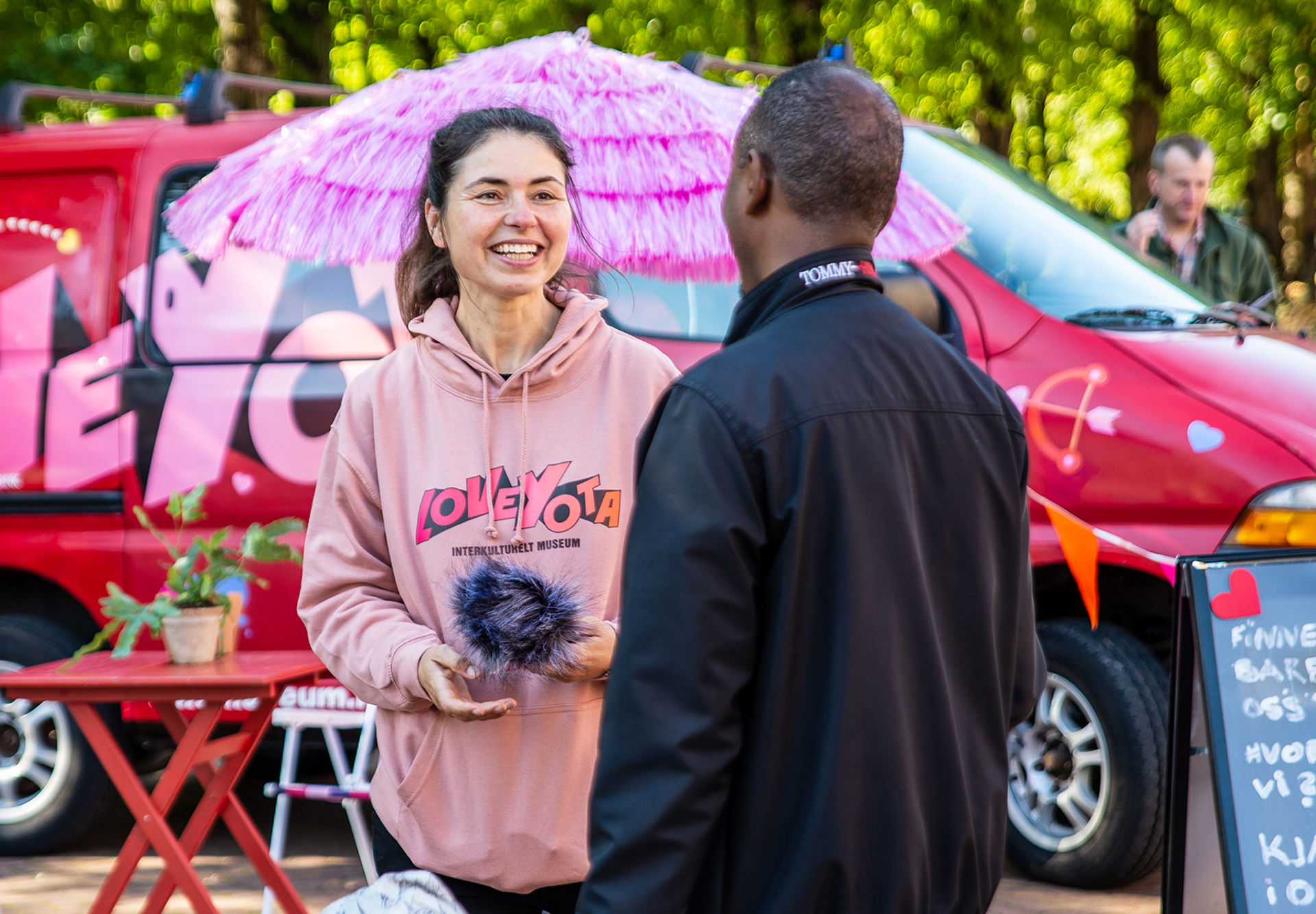 En person fra Oslo Museum intervjuer en mann som holder et barn i hånden. Fargerik bil i bakgrunnen.