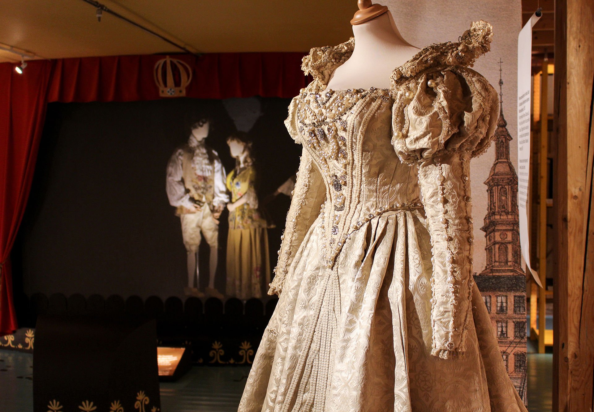 En flott kjole er hovedmotivet. I bakgrunnen to utstillingsdukker med teaterbekledning fra gamle dager.
