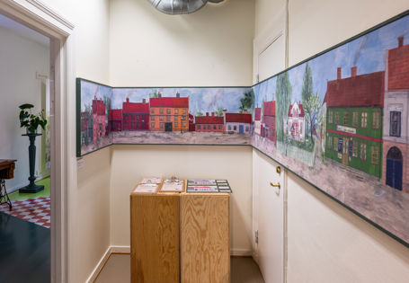 Hvitt lite rom med to trekasser med oppgaveark i midten. En stor avlang illustrasjon av gamle fargerike trehus går over alle tre veggene.