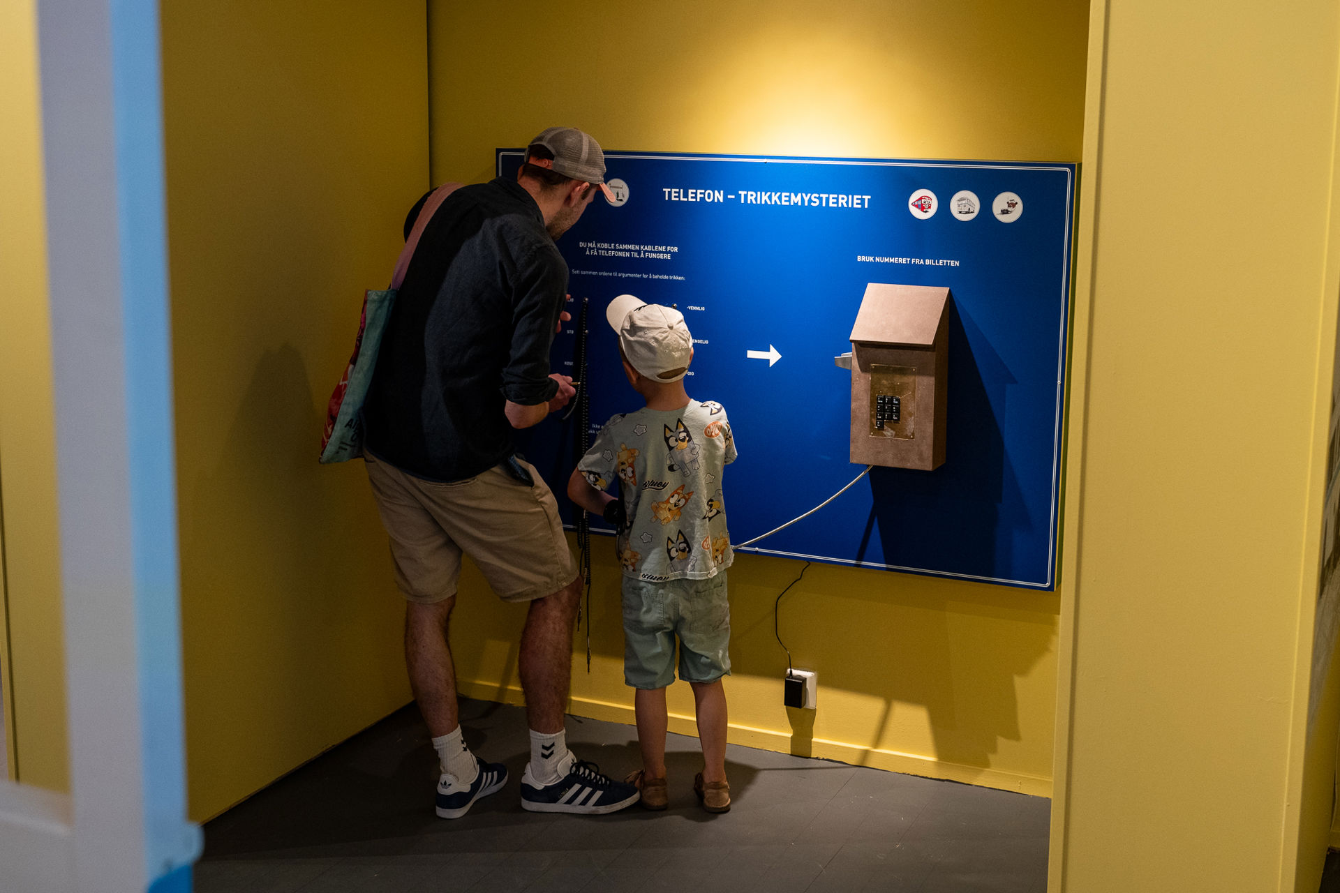 Mann og gutt i sommerlige klær med ryggen til. Står i et gult rom foran en blå tavle med en gammel telefon og teksten telefon trikkemysteriet.