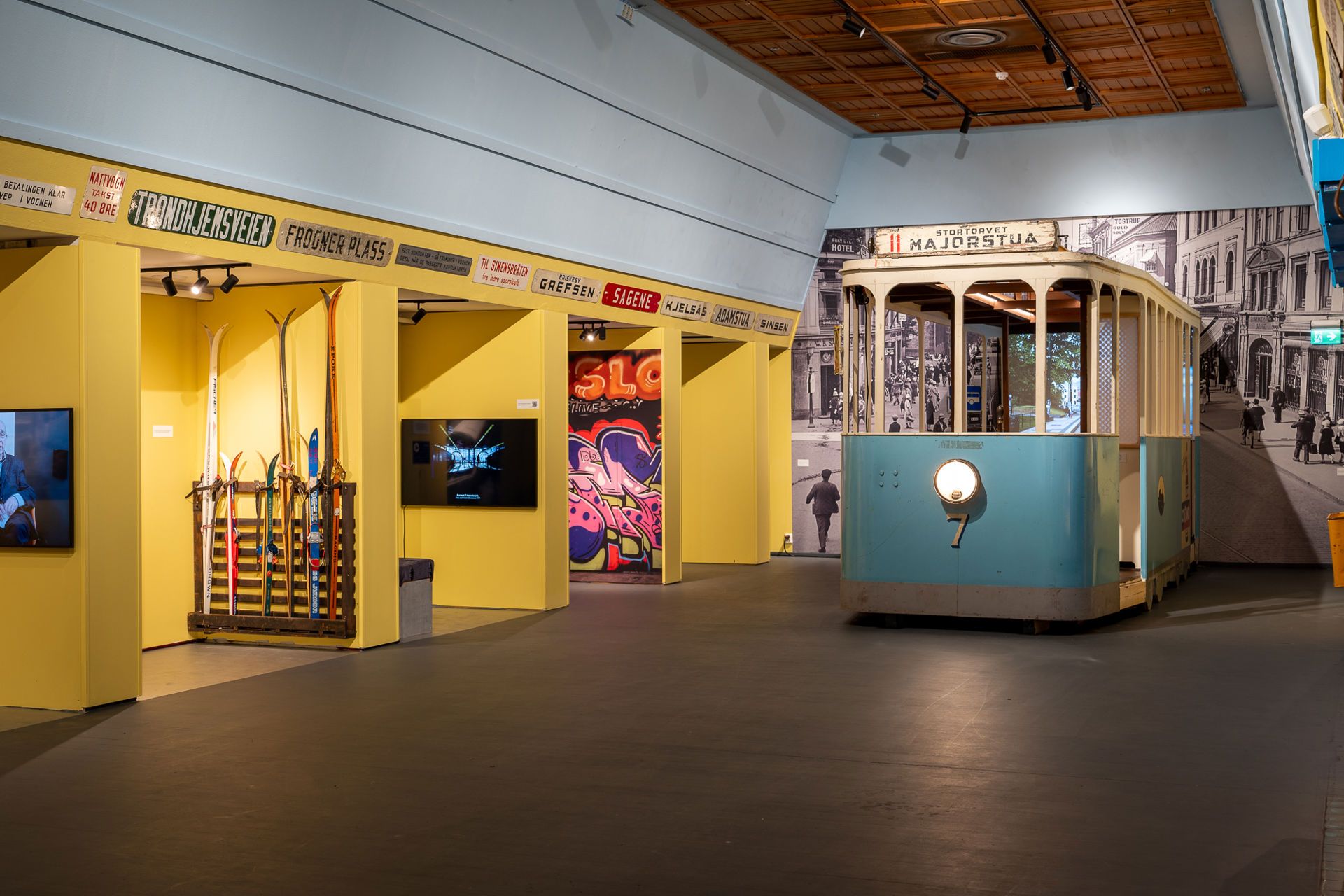En gammel blå trikkemodell stikker ut fra en vegg med et stort sort-hvitt-bybilde bak. Gule vegger rundt med skilt med ulike stedsnavn i Oslo.