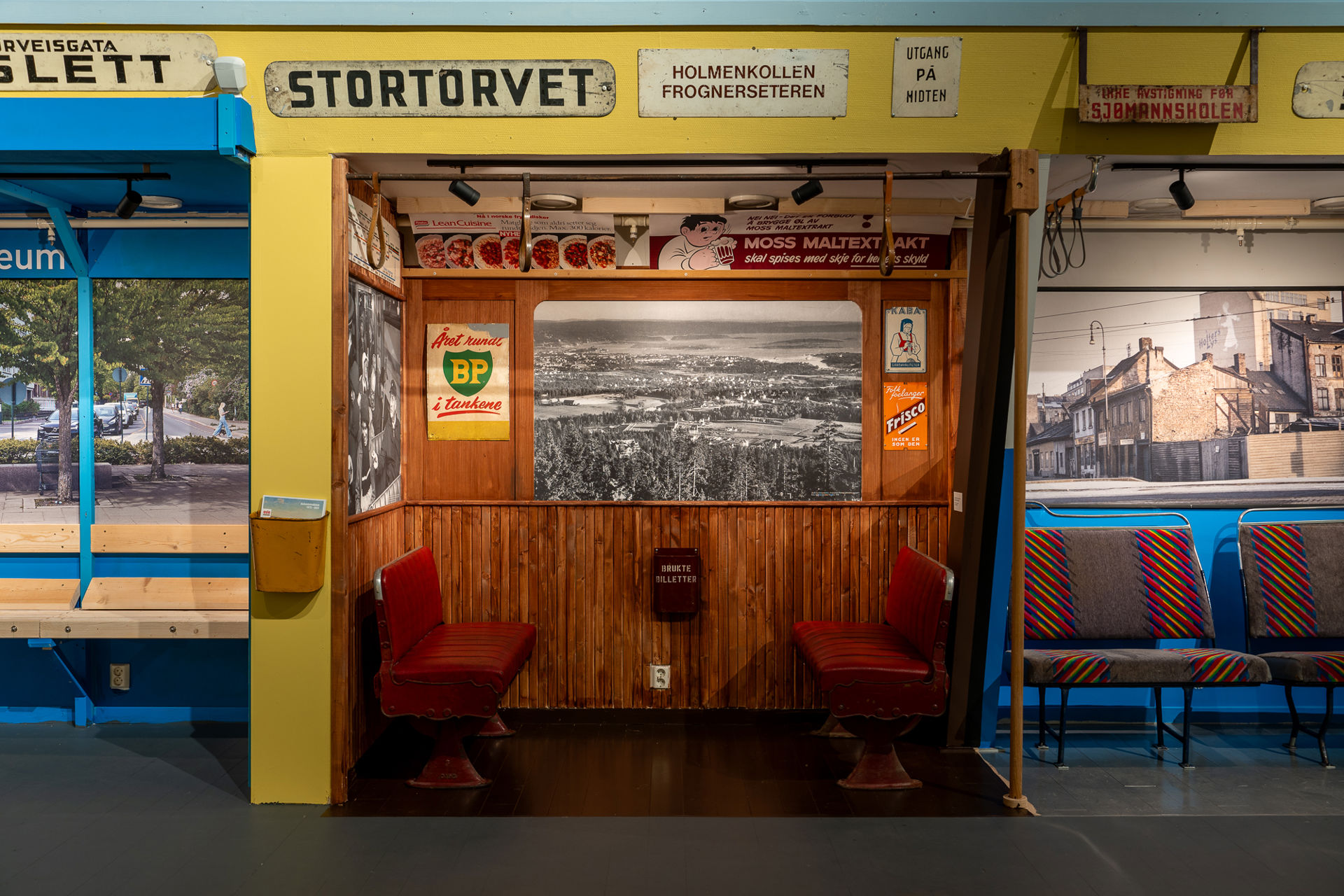 Replika av en gammel togvogn med trevegger og røde seter vendt mot hverandre, som del av en utstilling. Skilt med Stortorvet og Holmenkollen over vognen.