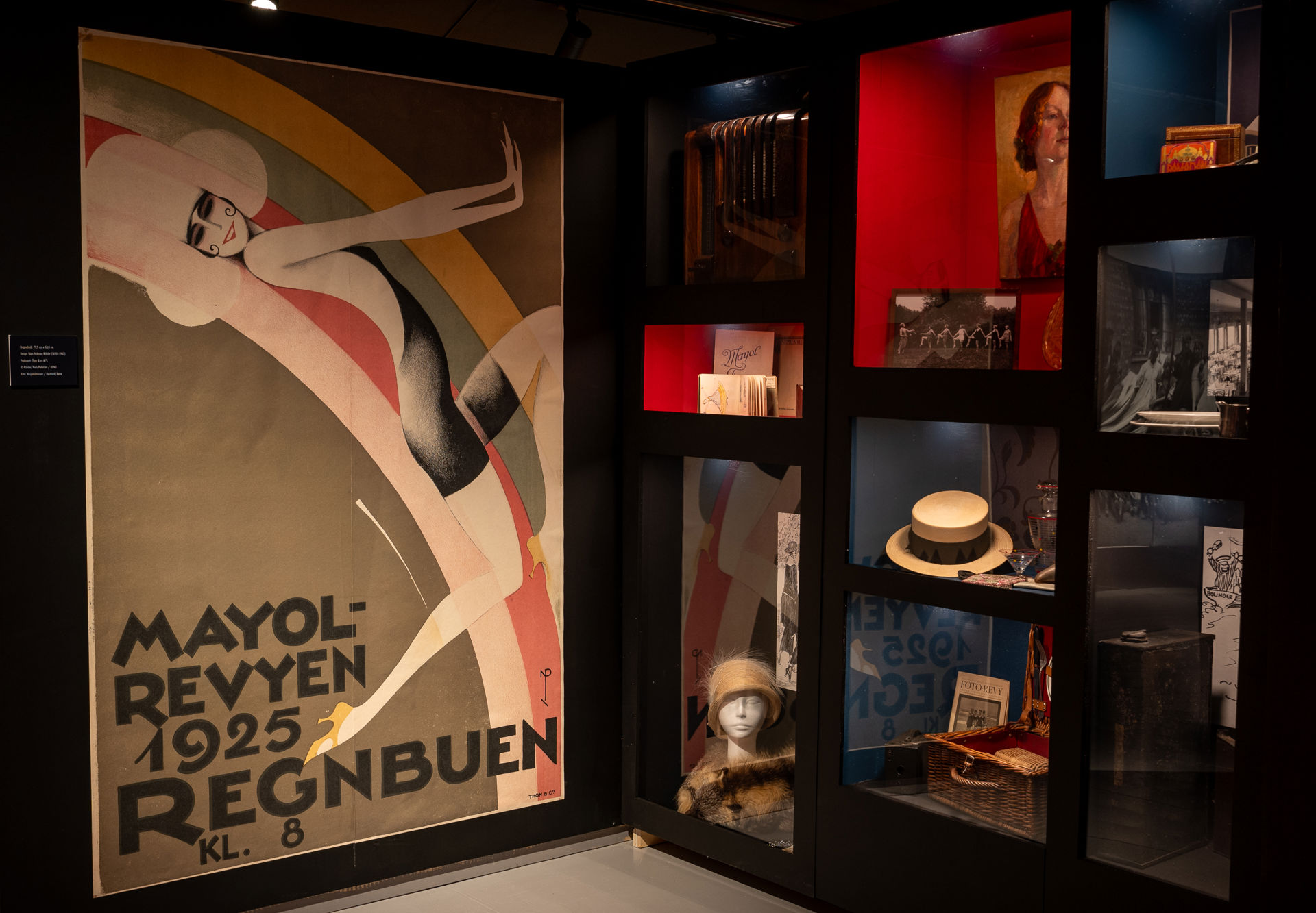 Stor plakat med illustrert kvinne og teksten Mayol-revyen 1925 Regnbuen kl. 8. Glassmontere med gjenstander til høyre.