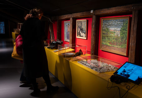 To personer med ryggen til ser ned på gjenstander i utstillingsmonter. Malerier på rød vegg foran dem