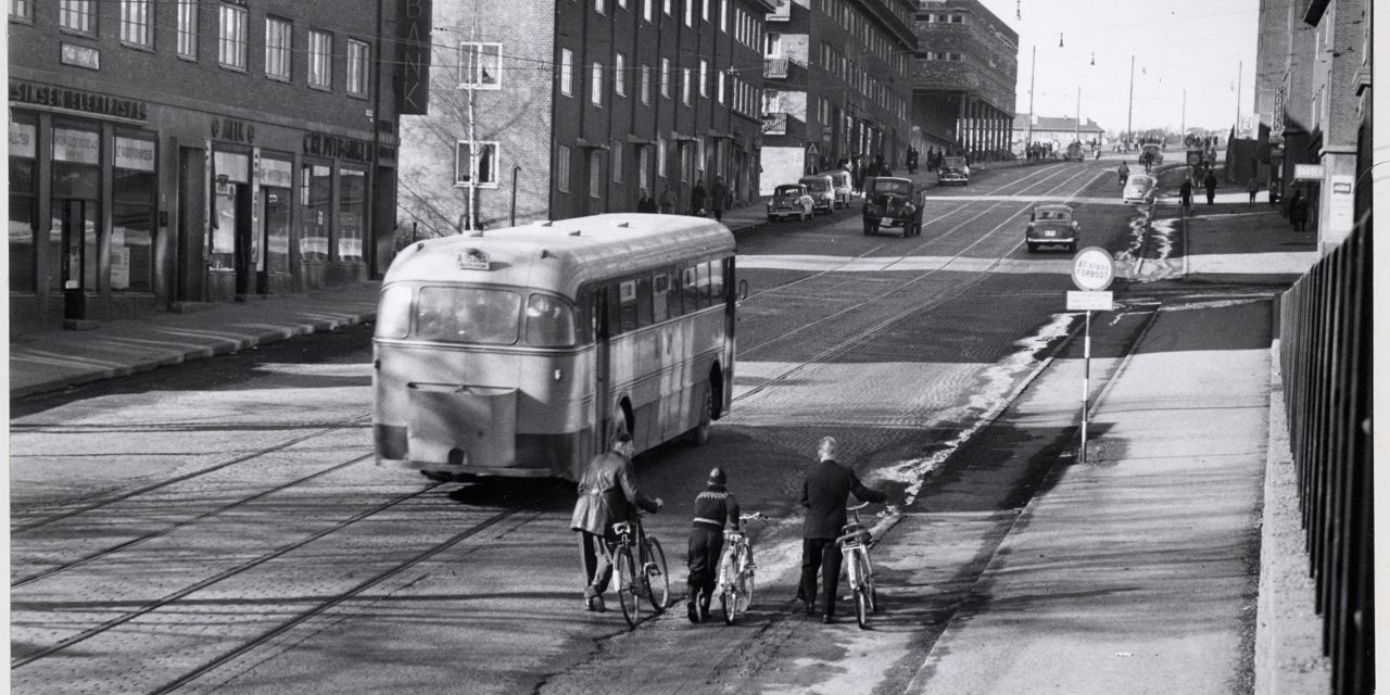Sort-hvitt-foto av en rett vei med trikkeskinner og blokker rundt. Gammeldags trikk og tre personer som triller sykler oppover veien i forgrunnen.