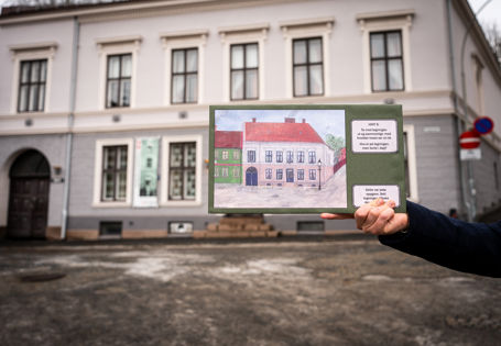 Hånd i forgrunnen holder en illustrasjon av et hus med spørsmål ved siden av. Samme huset sees i bakgrunnen.