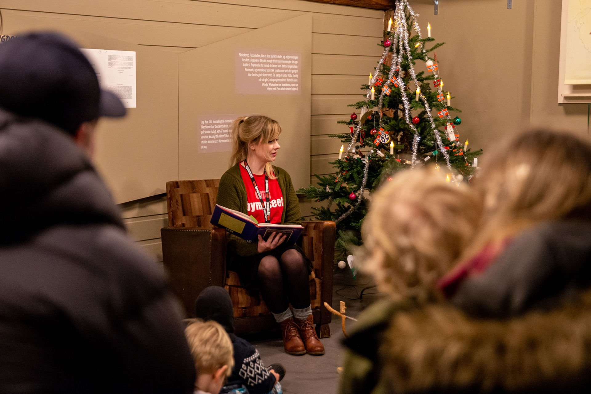 Kvinne sitter i en stol og leser eventyr for barn og voksne som står rundt henne. Et pyntet juletre står ved siden av.