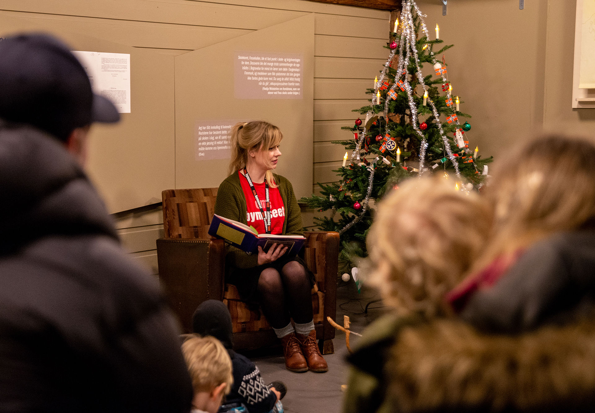 Kvinne sitter i en stol og leser eventyr for barn og voksne som står rundt henne. Et pyntet juletre står ved siden av.