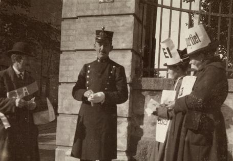 Tre personer står utenfor en bygning, med en uniformert politimann i midten og personer med plakater på hver side