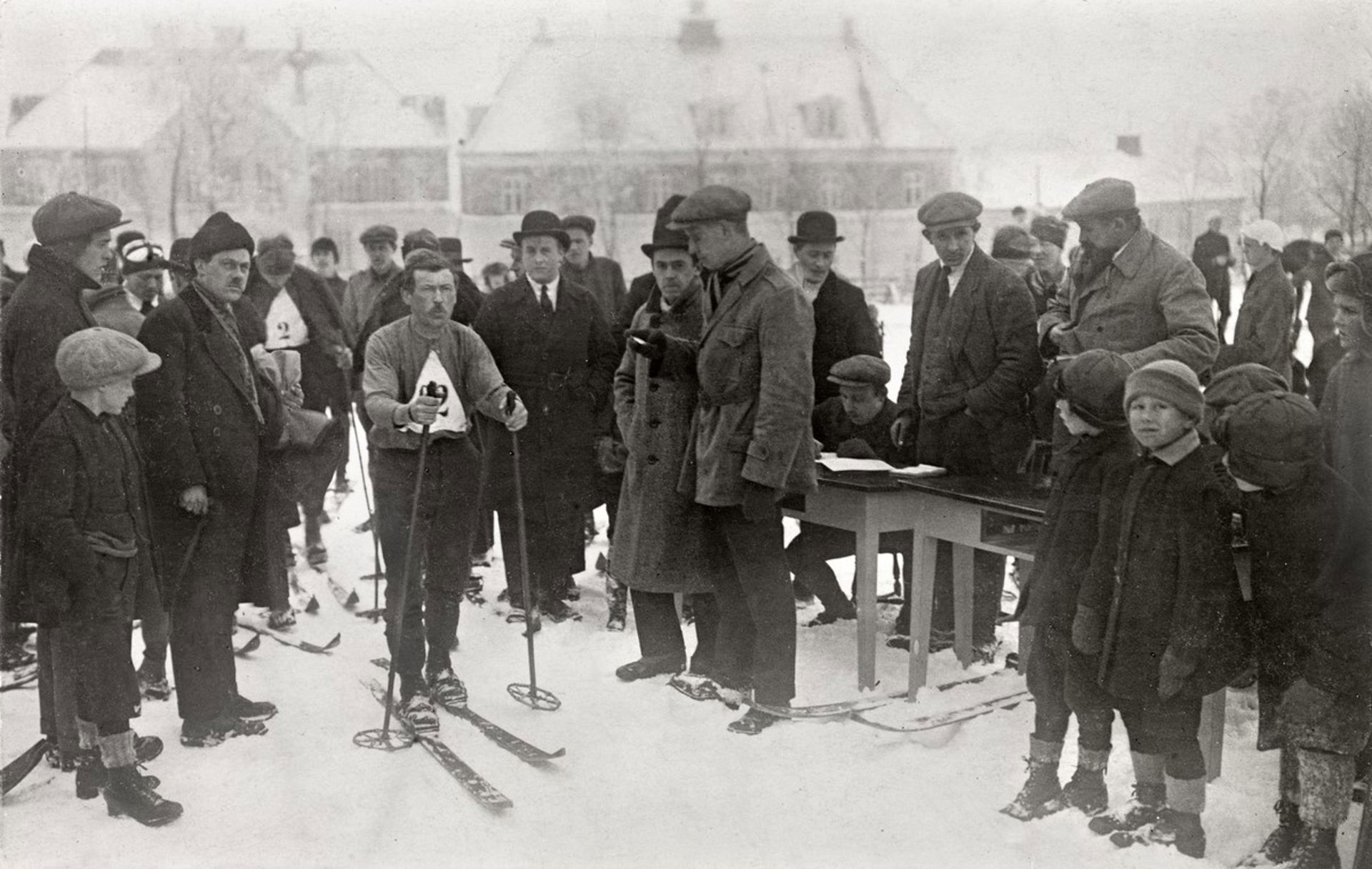 Mann med ski på beina og startnummer på brystet er omringet av mange mennesker i vinterklær.