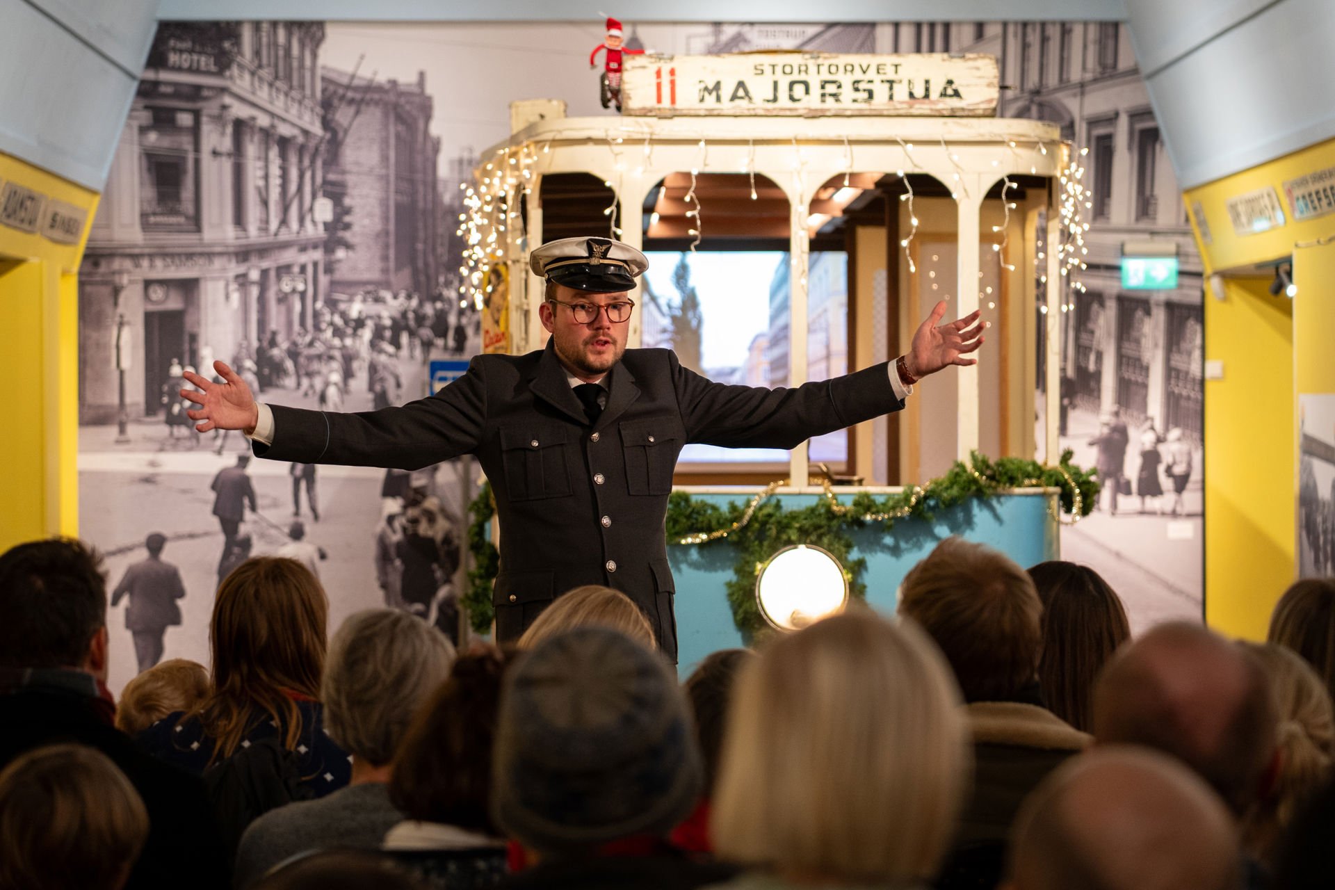 Mann i trikkeuniform med hvit lue og sort dress står med armene ut foran en blå trikk i en utstilling