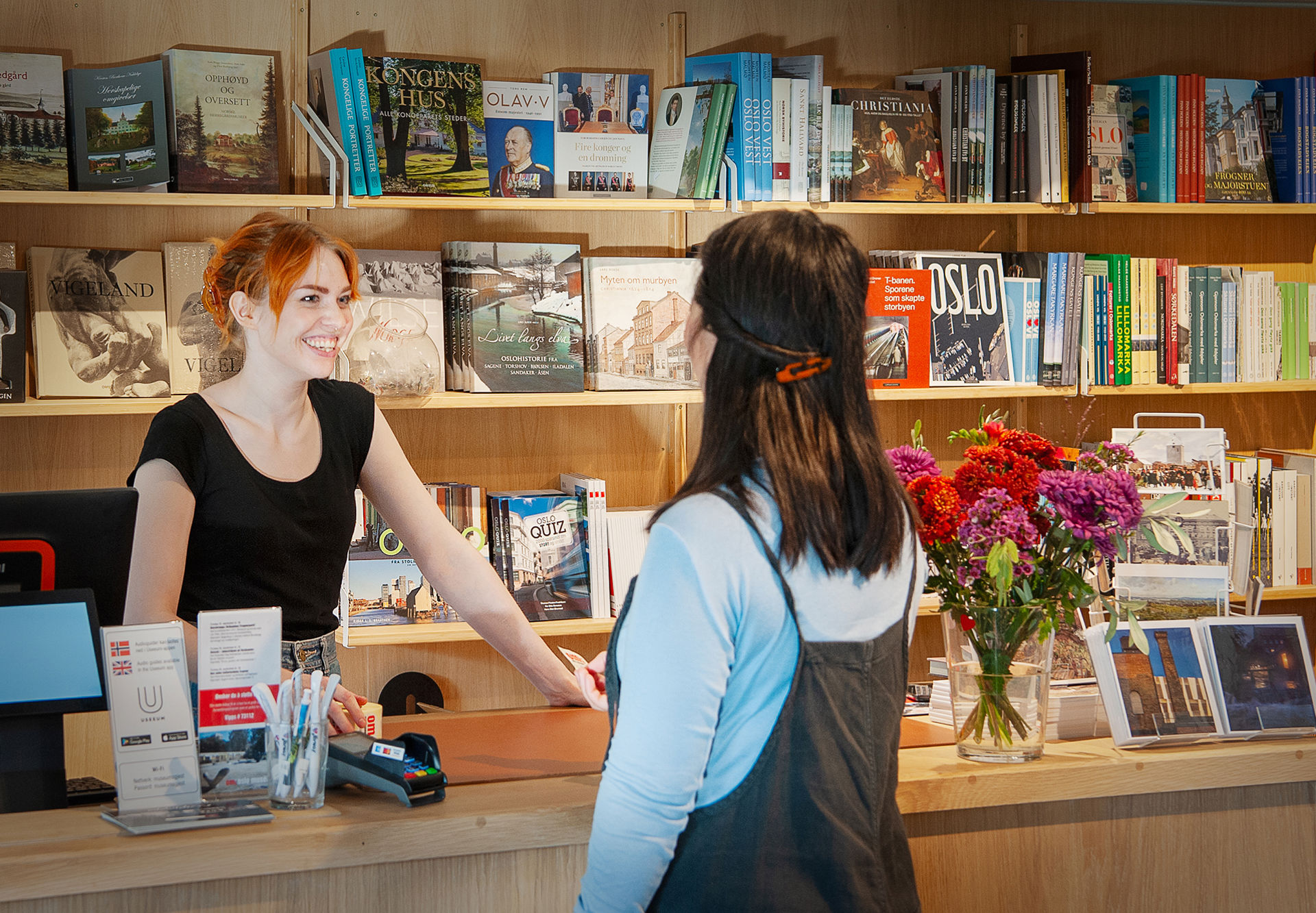 Ung kvinne bak kassen betjener kvinnelig kunde i butikken. Masse bøker i bakgrunnen.