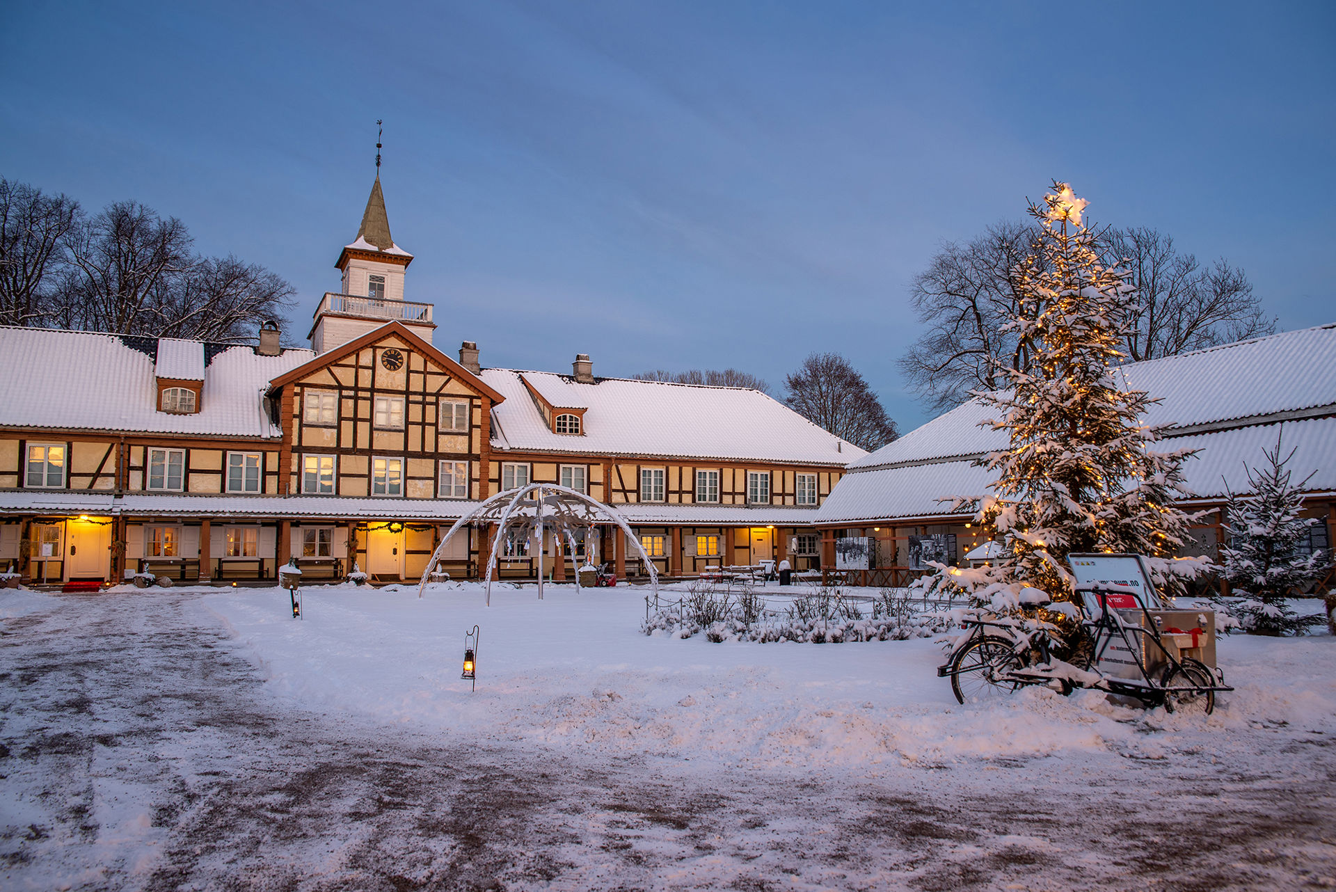Snø, juletre og gammelt stort bygg i fint kveldslys.