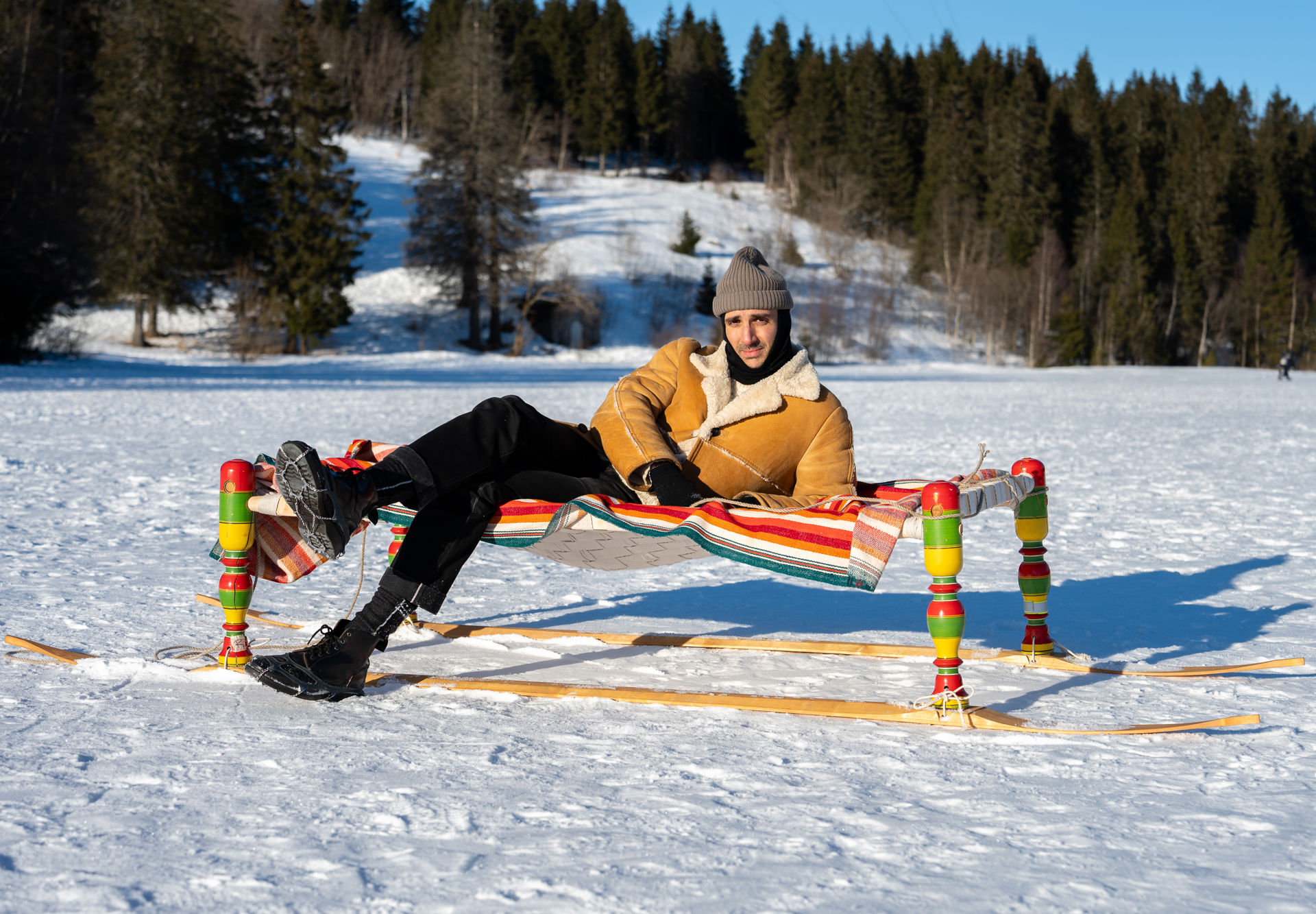 Mann i saueskinnsjakke ser mot kamera mens han ligger sidelengs på en dagseng med fargerike bein og treski montert på beina i snøen