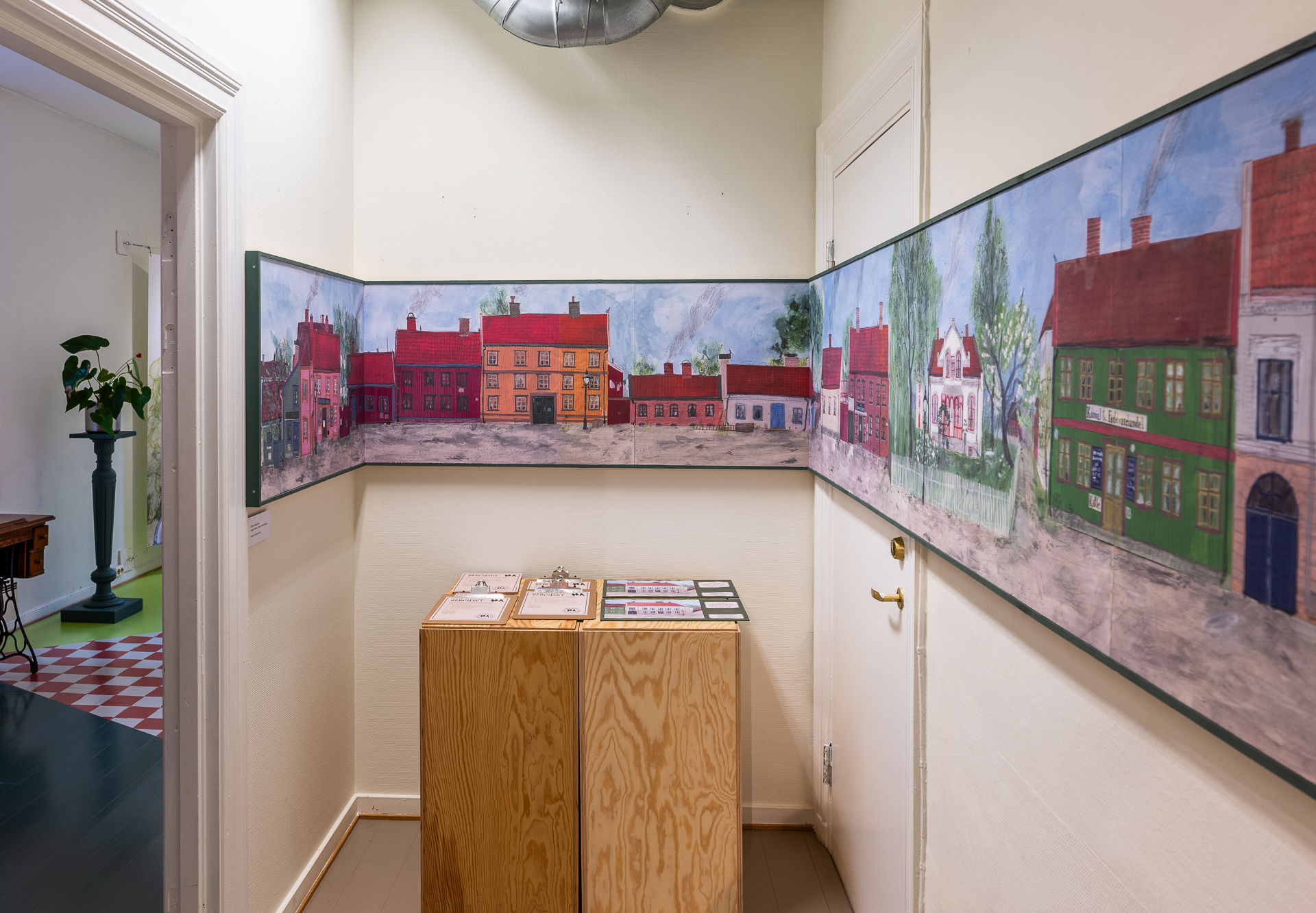 Hvitt lite rom med to trekasser med oppgaveark i midten. En stor avlang illustrasjon av gamle fargerike trehus går over alle tre veggene.