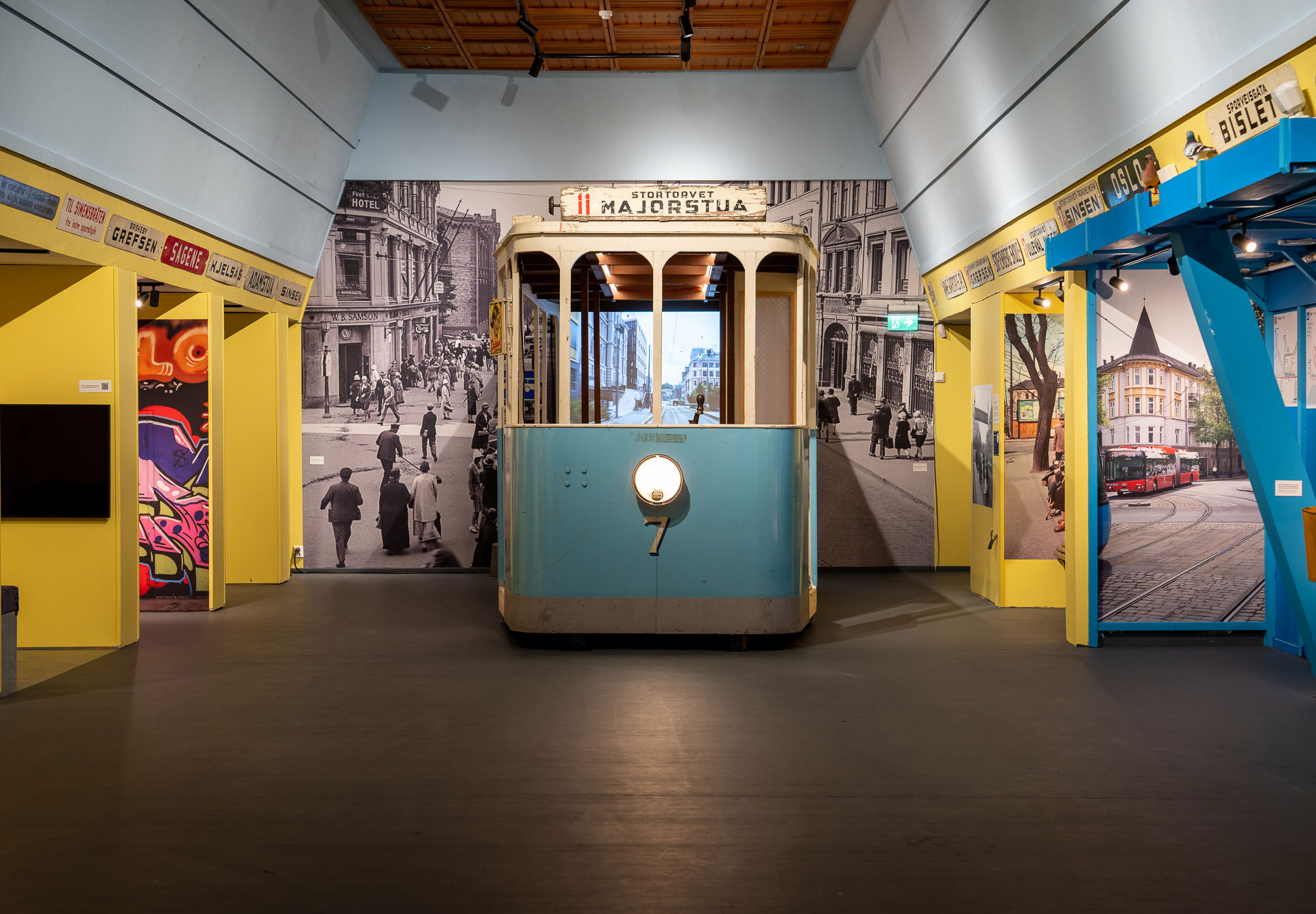 En gammel blå trikkemodell plassert midt i rommet med et sort-hvitt-bybilde bak. Gule vegger rundt med skilt med ulike stedsnavn i Oslo.