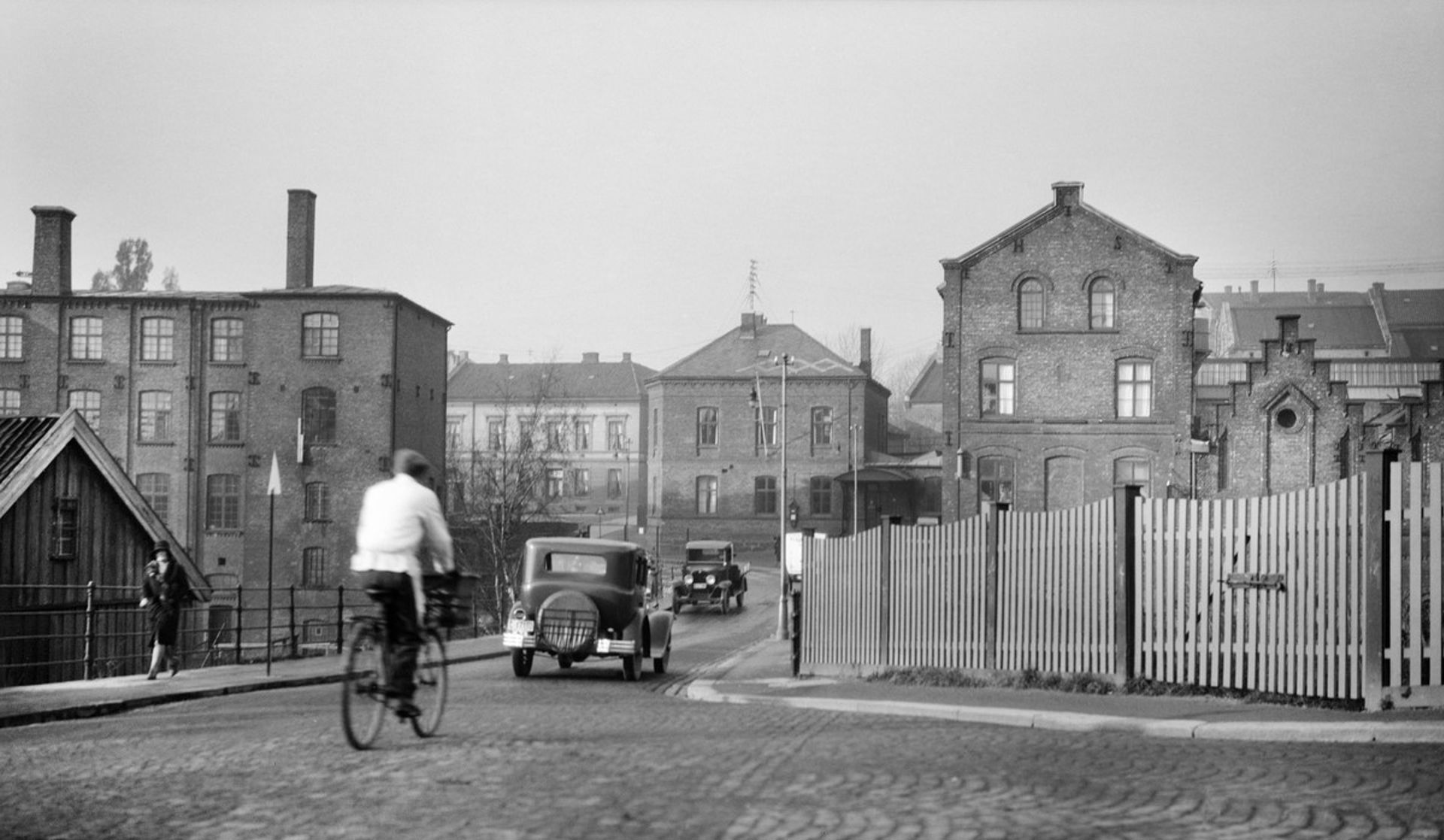 Sort-hvitt-bilde av mann som sykler etter en gammel bil over en bru. Fabrikkbygninger på hver side av brua.