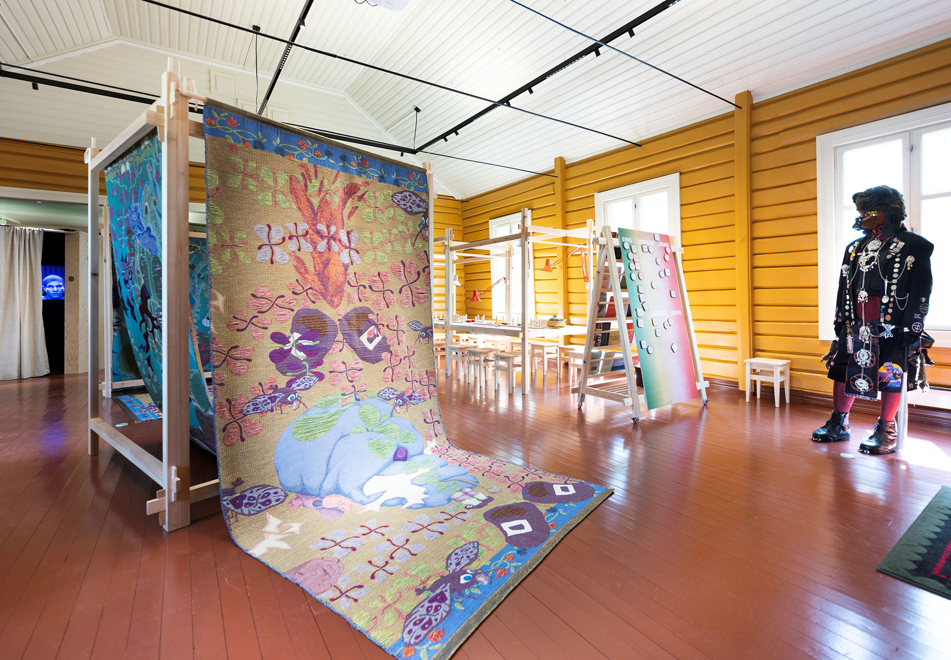 Fargerike tekstilinstallasjoner hengt opp i en trerigg, en menneskelignende figur kledd i sort med symboler og merker på klærne til høyre