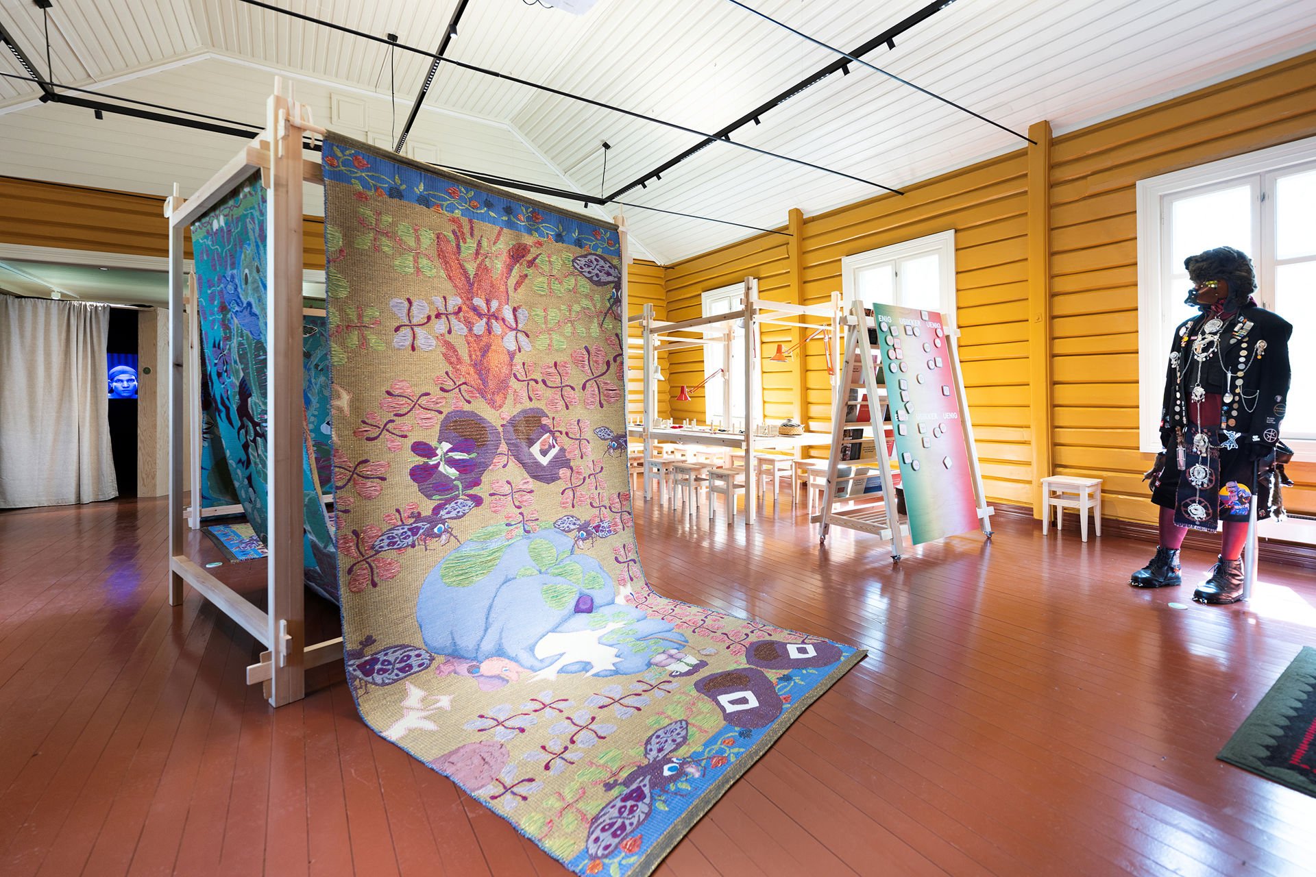 Fargerike tekstilinstallasjoner hengt opp i en trerigg, en menneskelignende figur kledd i sort med symboler og merker på klærne til høyre